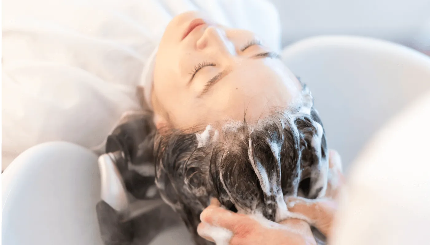 美容院でシャンプーを受ける女性。リラックスした表情で目を閉じ、泡立てたシャンプーで丁寧に髪を洗ってもらっています。